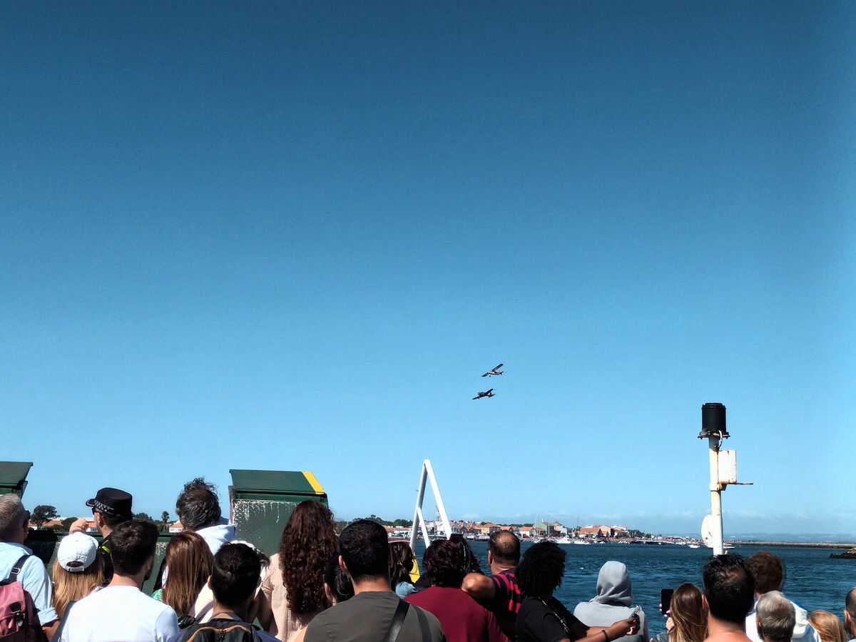 The photo shows a crowd of onlookers aboard the old ferry watching a Fly-by at the Dunas Festival Airshow in 2023, with the fishing harbor of São Jacinto in the background 