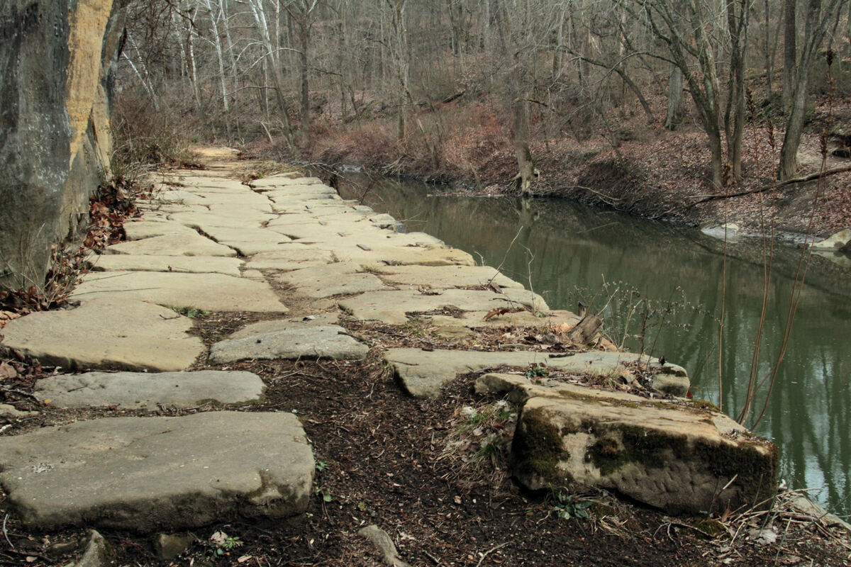 A pathway made of large, flat stones runs alongside a calm river. Bare trees and leafless branches are visible on both sides.